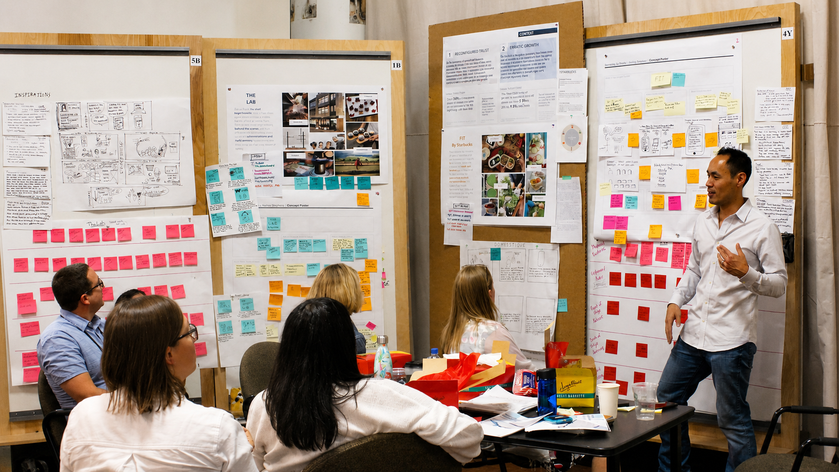 Andrew facilitating a Discover + Design workshop at Starbucks, standing at a wall of concept posters and sticky notes with seated participants in the foreground.